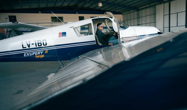 Pilot in a Piper aircraft with hangar setting, ready for takeoff.